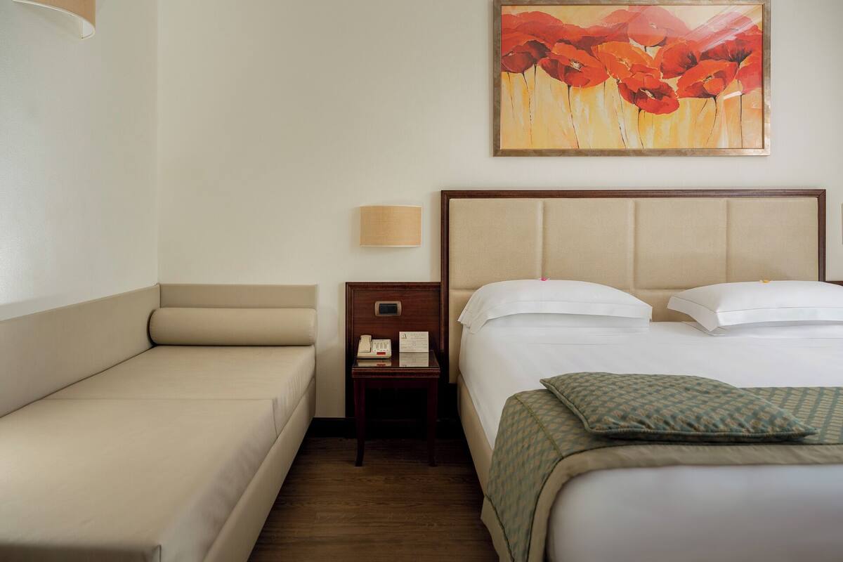 A hotel room with a bed, a sofa, a nightstand, a lamp, and a painting on the wall.