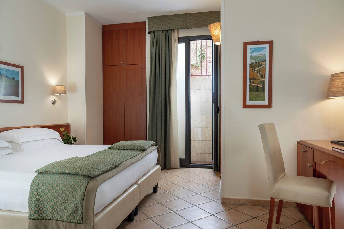 A hotel room with a bed, a desk, a chair, a wardrobe, a door leading to a balcony, and a wall-mounted lamp.