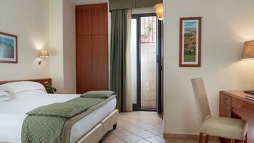 Habitación de hotel con cama, escritorio, silla, armario, puerta que da a un balcón y una lámpara de pared.