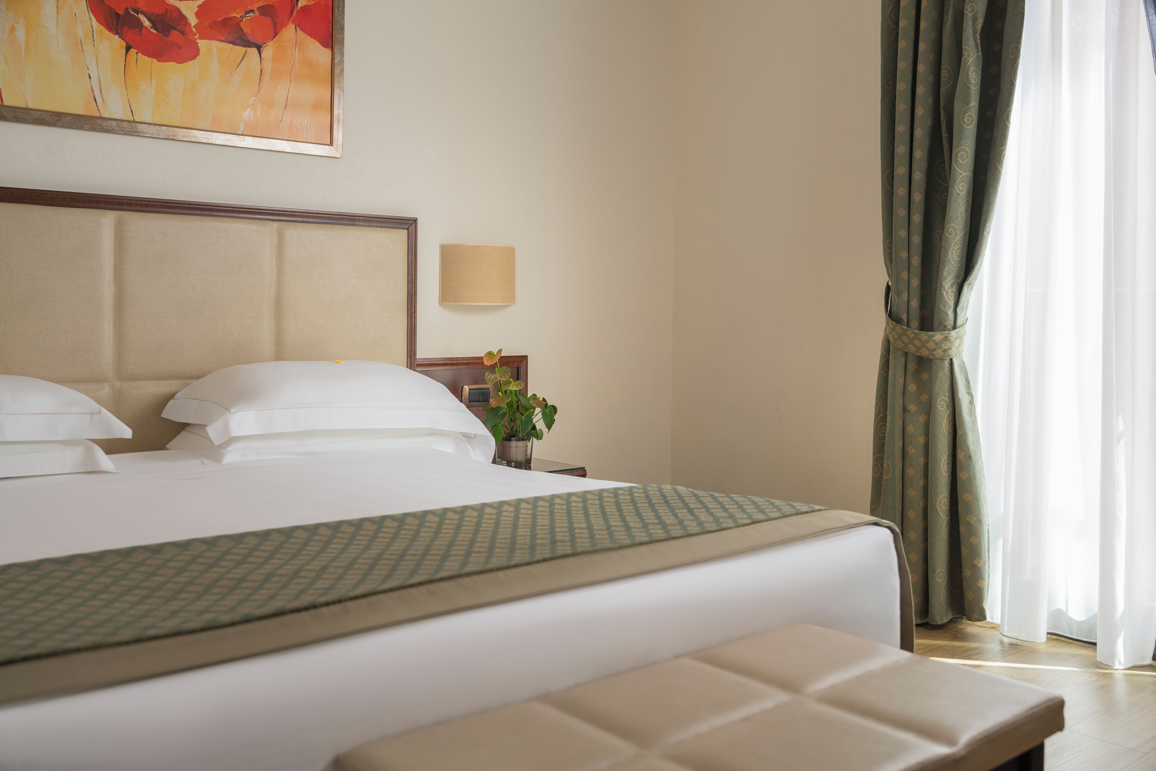 A hotel room with a bed, a bedside table with a lamp, a plant, and a painting on the wall.
