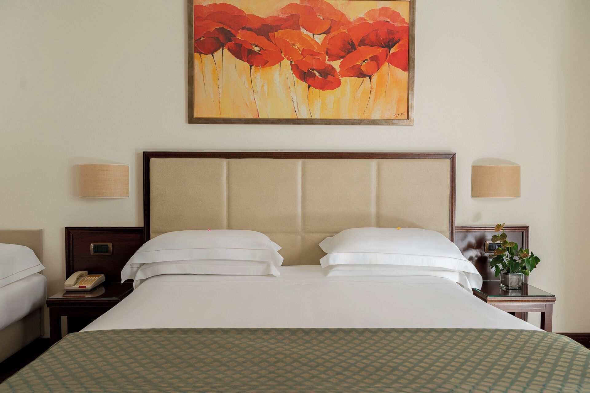 Ein Hotelzimmer mit einem großen Bett, zwei Nachttischen, einem Beistelltischchen mit Telefon und einem Gemälde von roten Blumen an der Wand.
