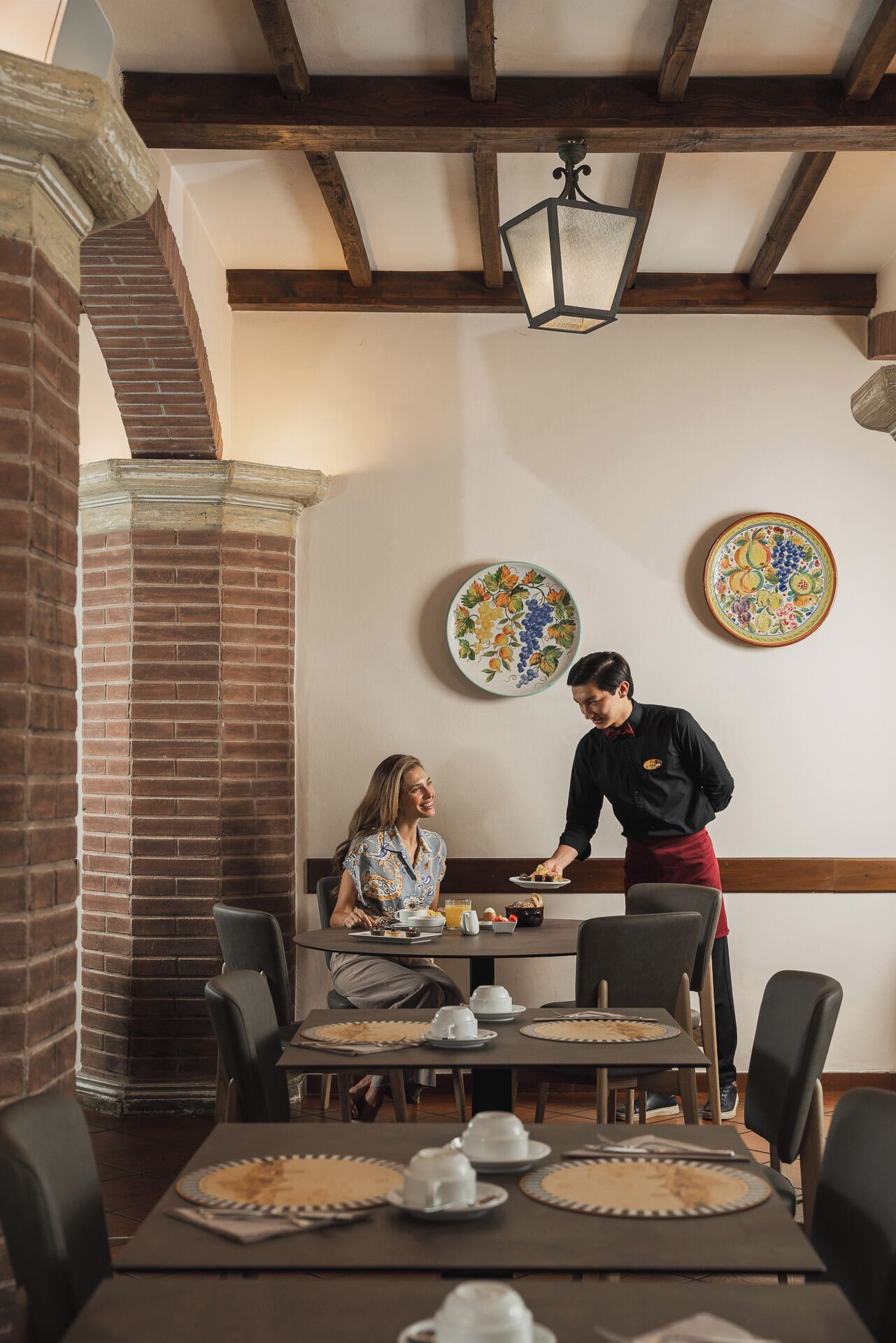 Un comedor con paredes de ladrillo, vigas de madera y chimenea de ladrillo. Hay dos mesas dispuestas para comer, con platos, vasos y un frutero.