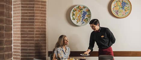 Un comedor con paredes de ladrillo, vigas de madera y chimenea de ladrillo. Hay dos mesas dispuestas para comer, con platos, vasos y un frutero.