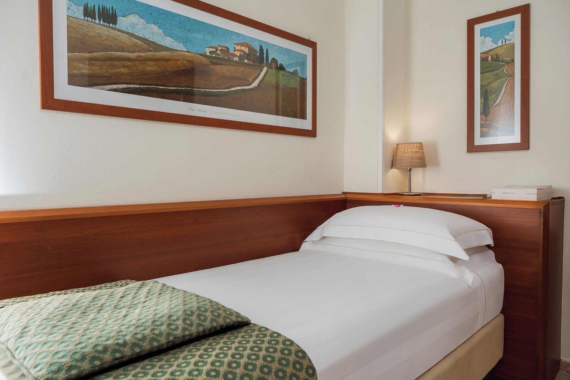 A hotel room with a bed, a nightstand, a lamp, and two framed pictures on the wall.