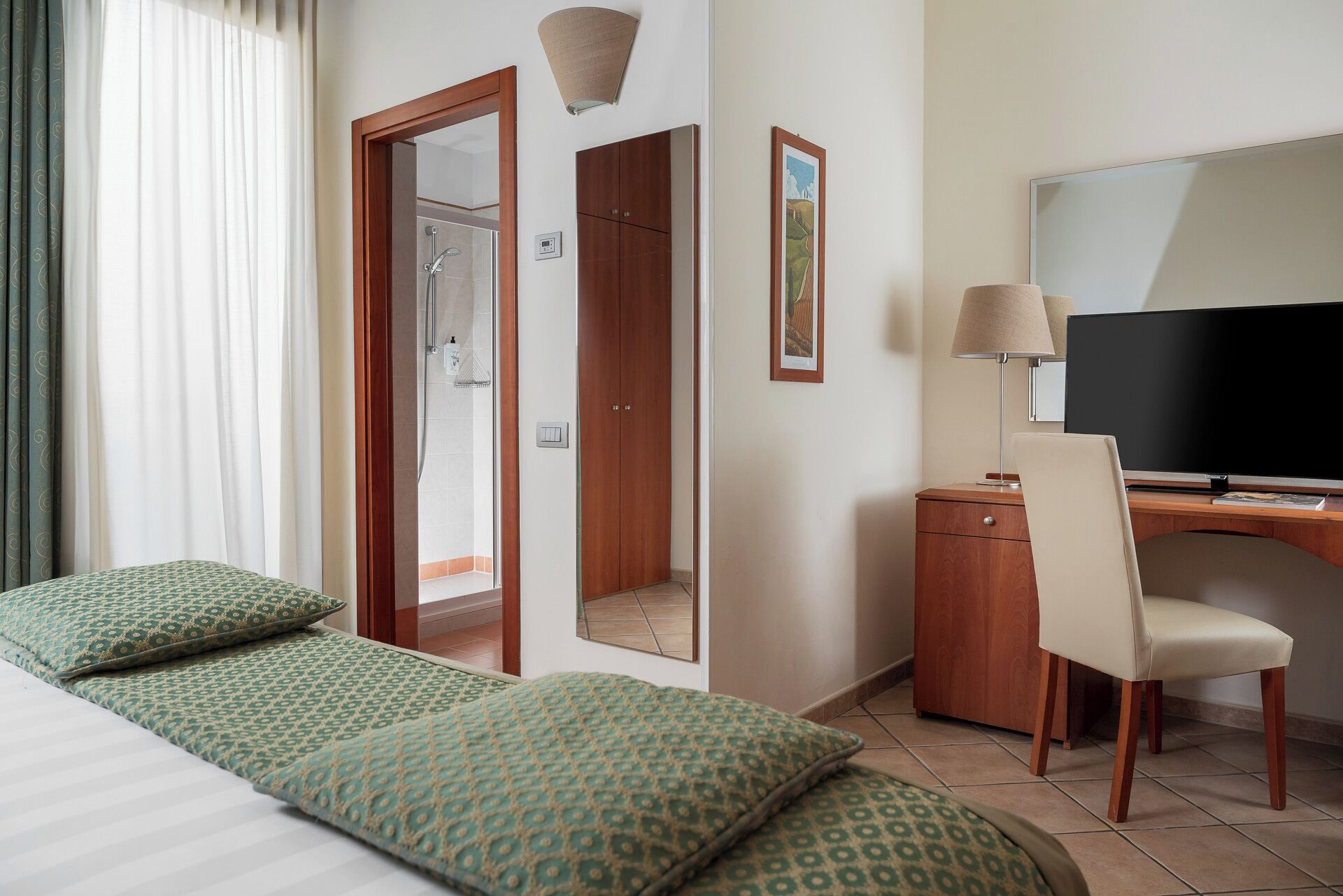 A hotel room with a bed, a desk, a chair, a TV, a shower, and a painting on the wall.