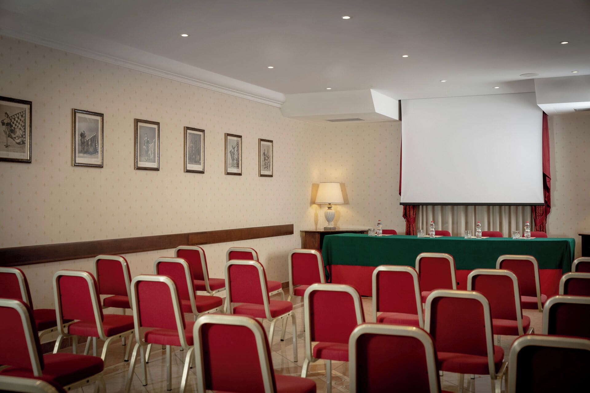 Ein Konferenzraum mit Reihen roter Stühle, einer Projektionsleinwand, einem Tisch mit grüner Tischdecke und gerahmten Bildern an der Wand.