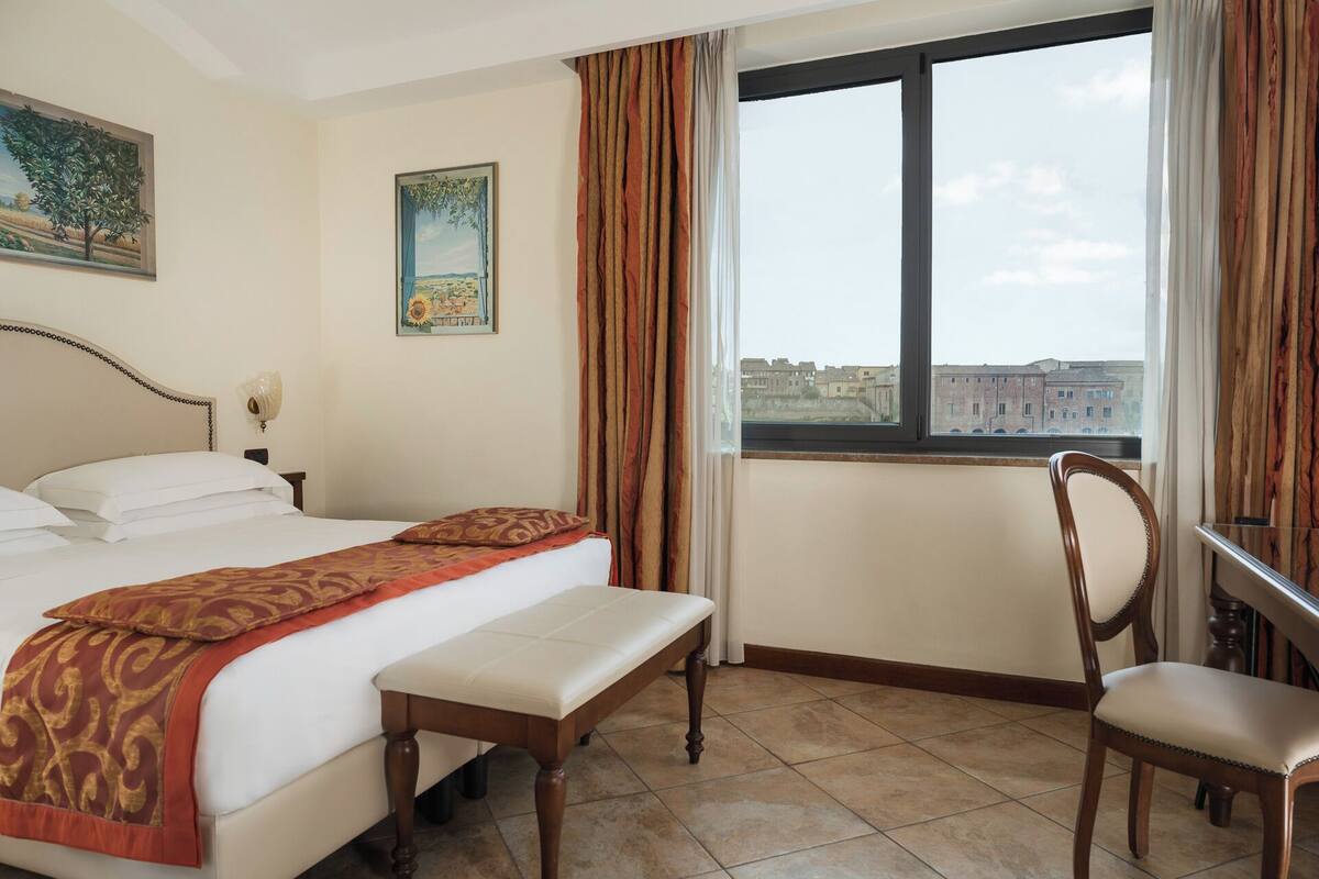 A hotel room with a bed, a bench, a chair, a window with a view of buildings, and two paintings on the wall.