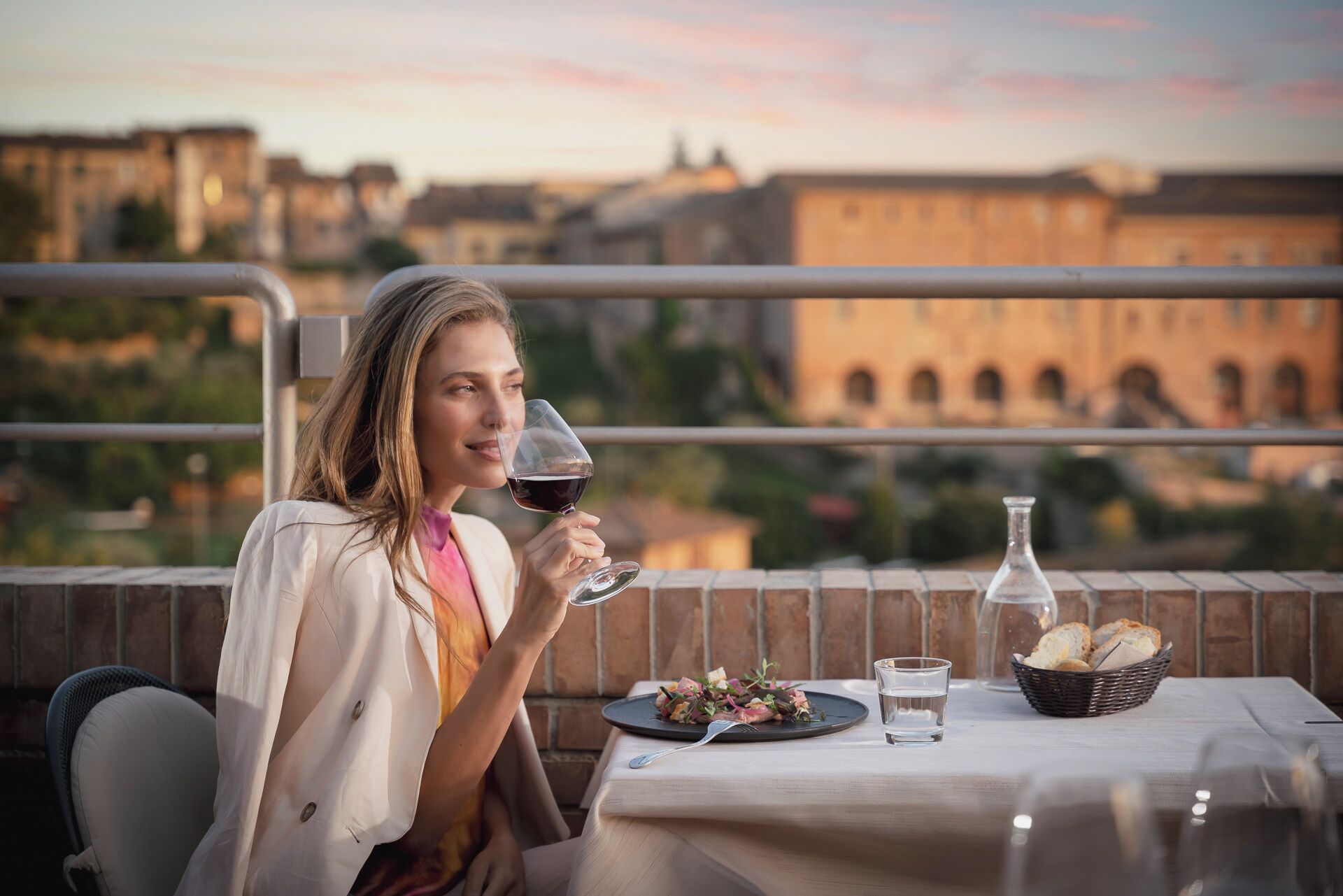 A person seated at a table with a meal, a glass of red wine, and a view of buildings in the background.