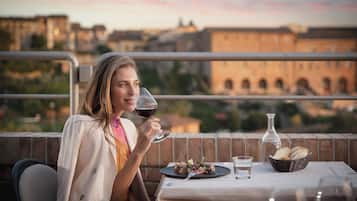 Une personne est assise à une table, devant un repas, avec un verre de vin rouge, et on aperçoit des bâtiments en arrière-plan.