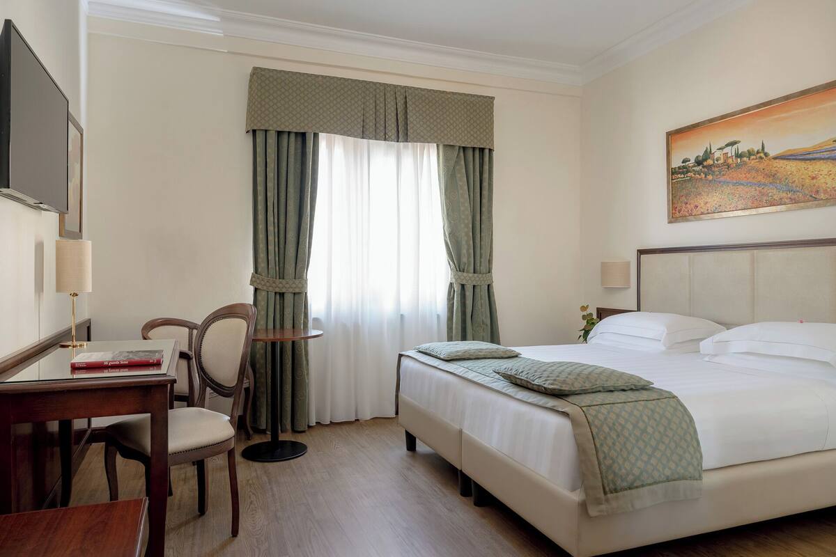 A hotel room with a bed, a desk with a chair, a lamp, and a painting on the wall.