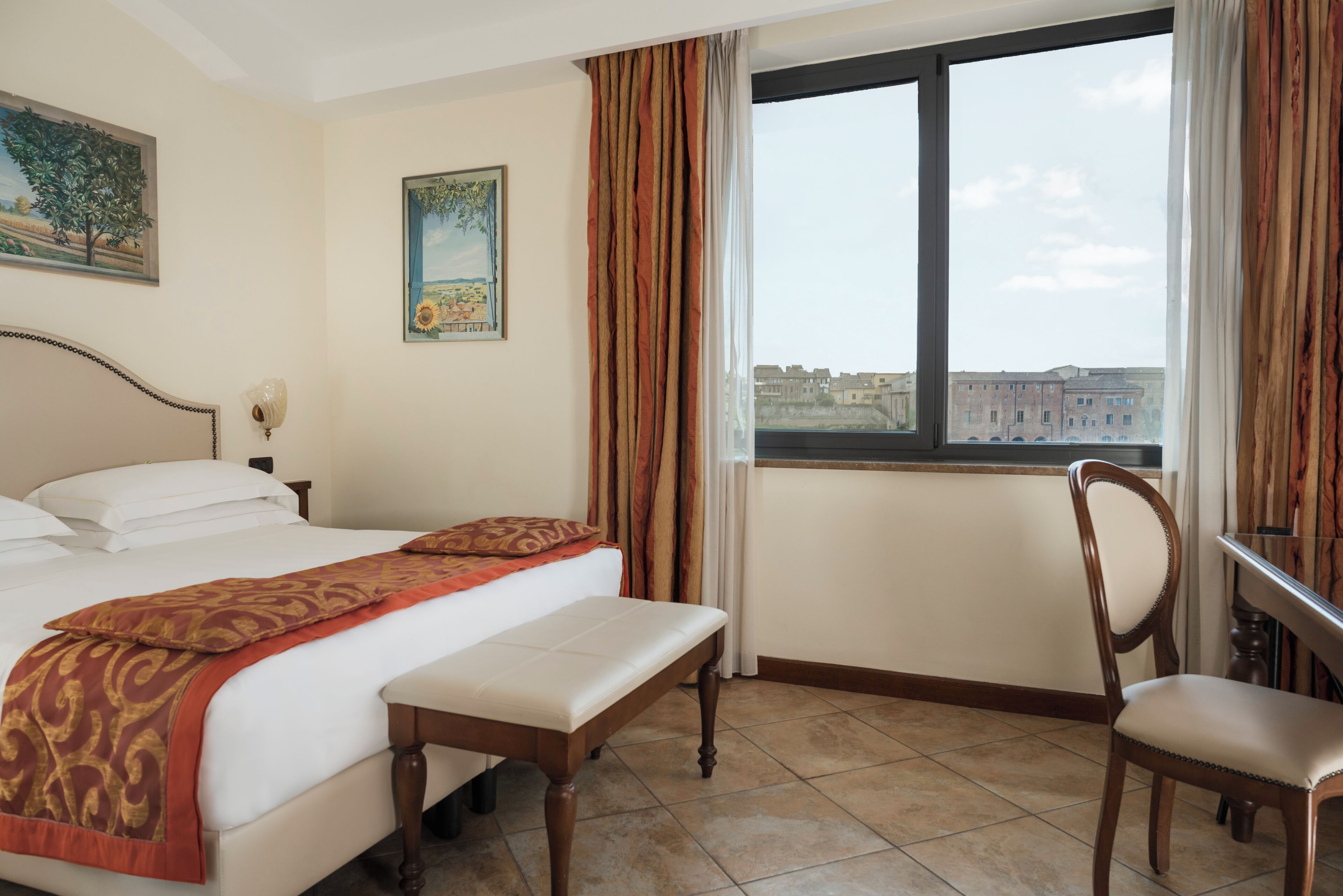 A hotel room with a bed, a bench, a chair, a desk, a window with a view, and two paintings on the wall.