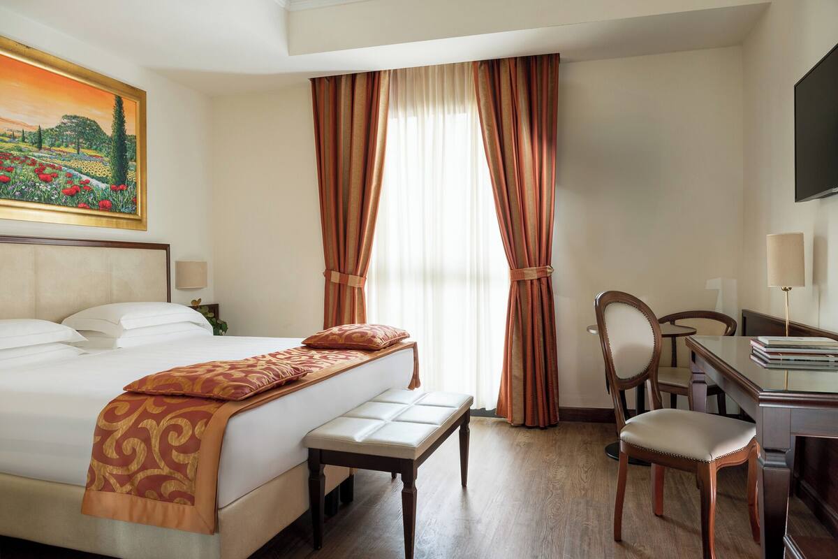 A hotel room with a bed, a desk, a chair, and a painting on the wall.