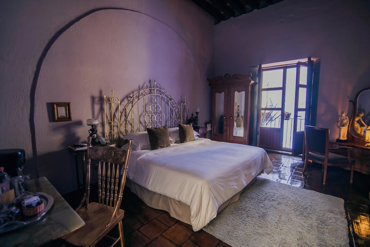 A bedroom with a large bed, a wooden chair, a small table with a tray, a mirror, and a window with a view of the outside.