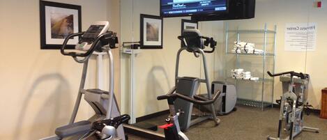 Un gimnasio con máquinas de cardio, un televisor mostrando un programa deportivo y un espejo que refleja el equipo.