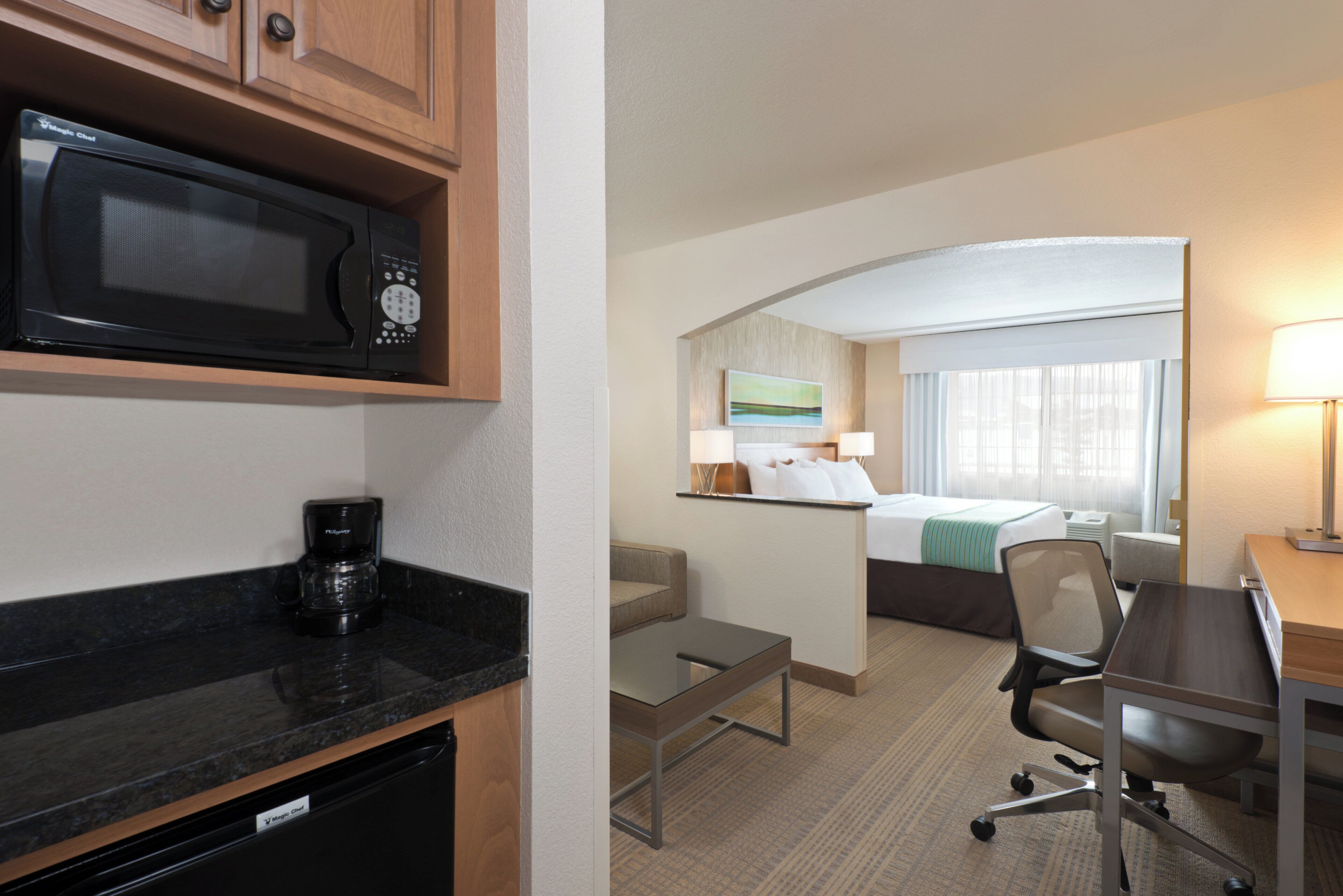 A hotel room with a bed, a desk, a chair, a coffee maker, a microwave, and a small table.