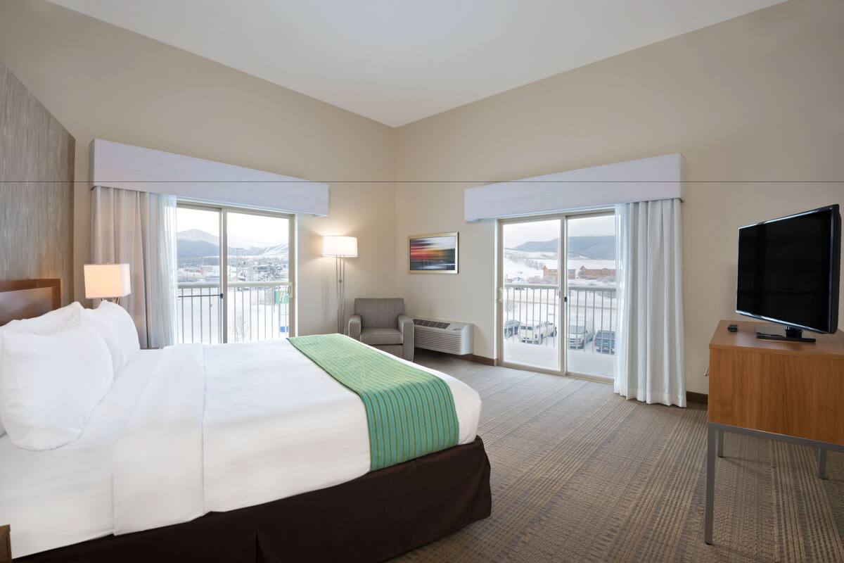 A hotel room with a large bed, a desk, a chair, a TV, and a view of the city through the windows.