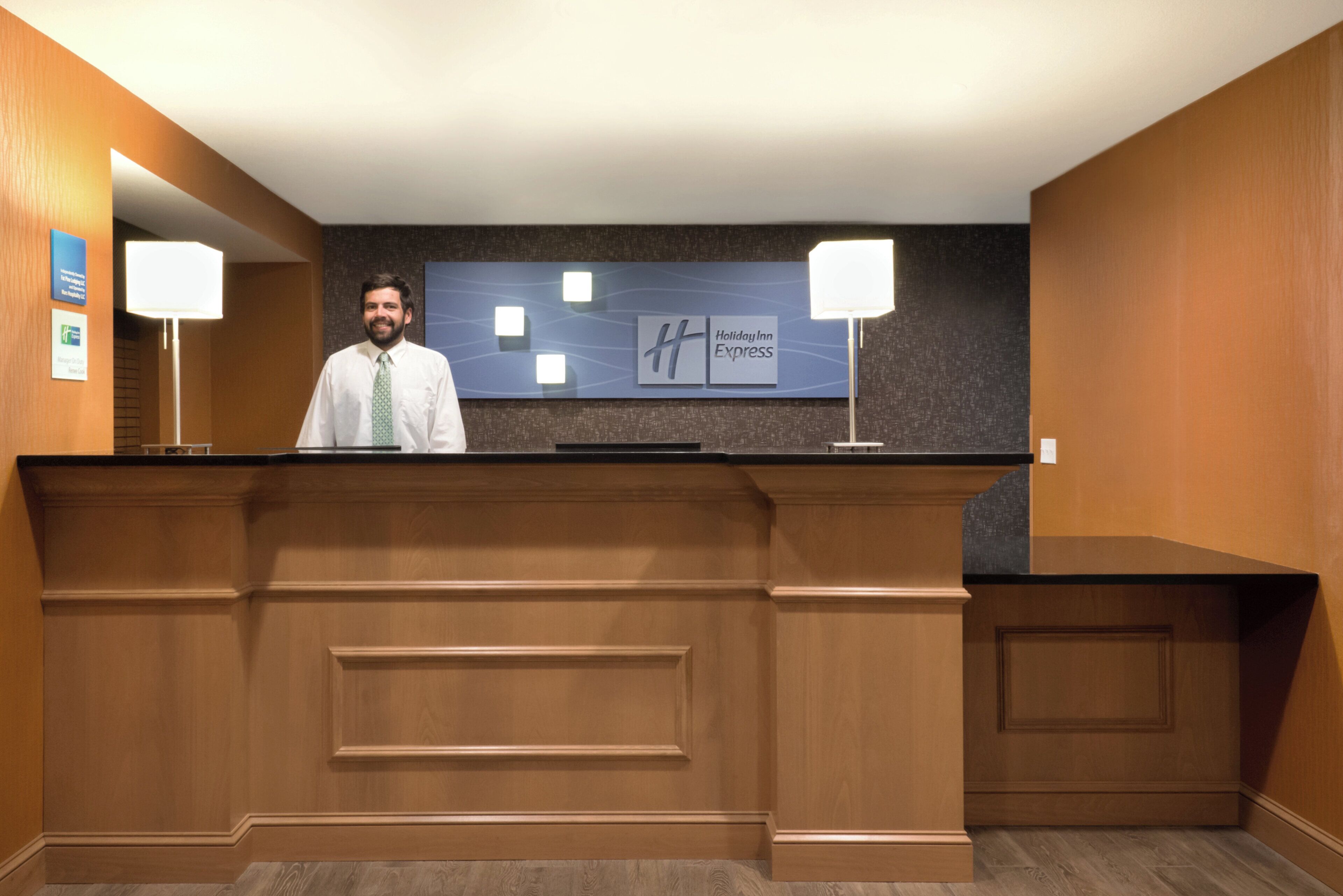 A hotel reception desk with a man standing behind it, a Holiday Inn Express sign above, and two wall-mounted lamps on either side.