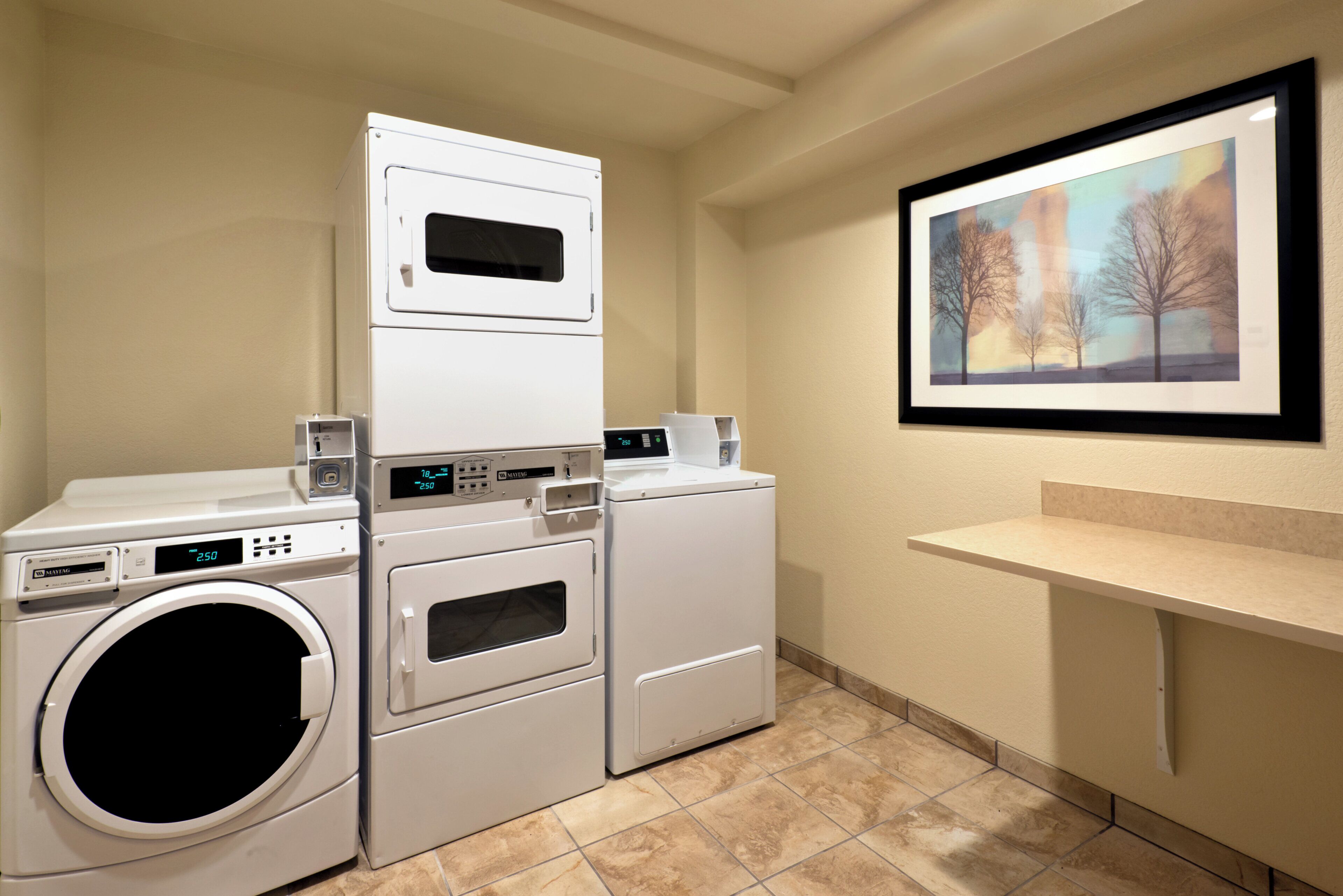 A laundry room with stacked washers and dryers, a digital display, and a framed landscape painting on the wall.