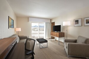 Habitación de hotel con escritorio, dos sillas, televisor, lámpara y vistas a montañas a través de la ventana.