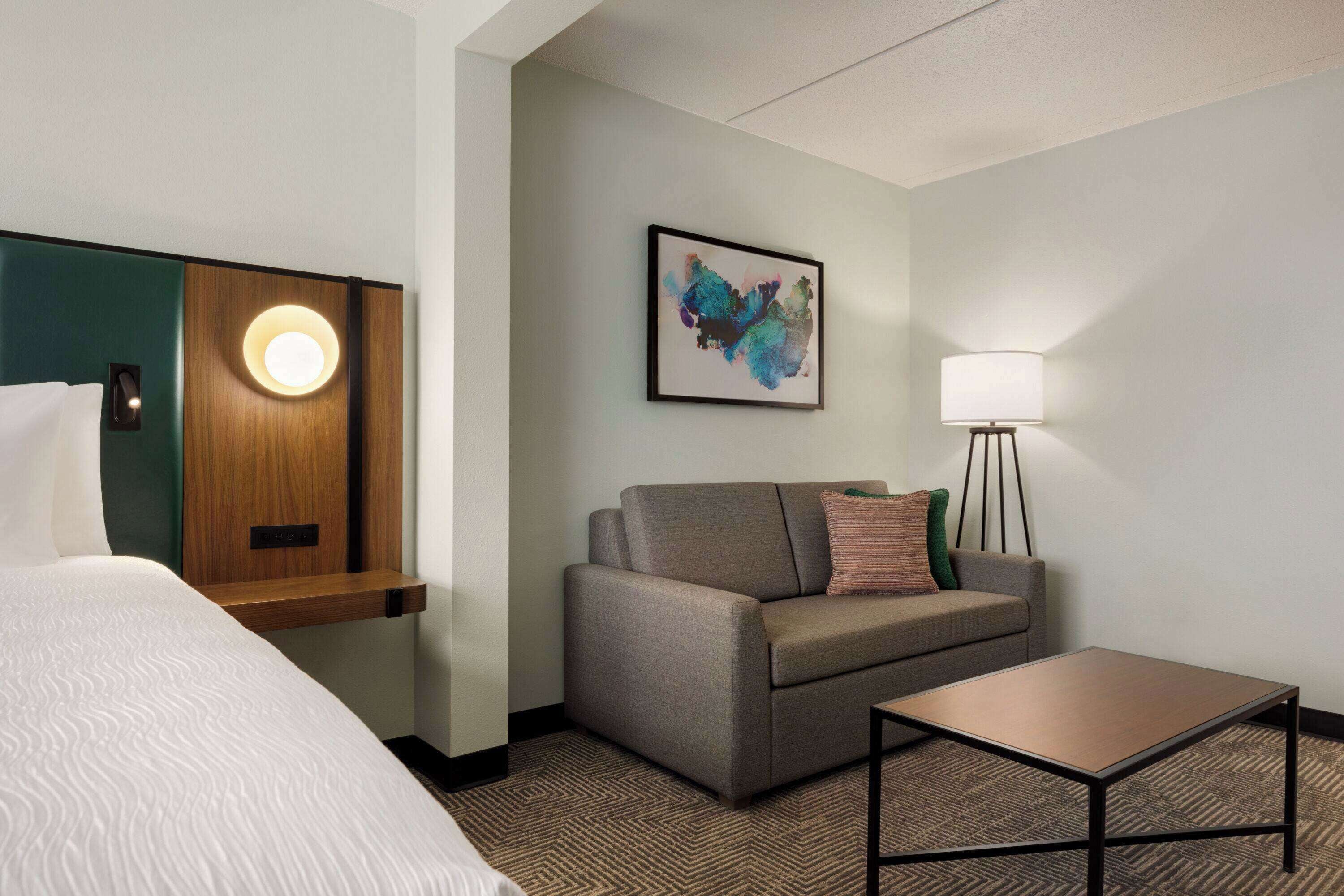 A hotel room with a bed, a sofa, a small table, a lamp, and a painting on the wall.