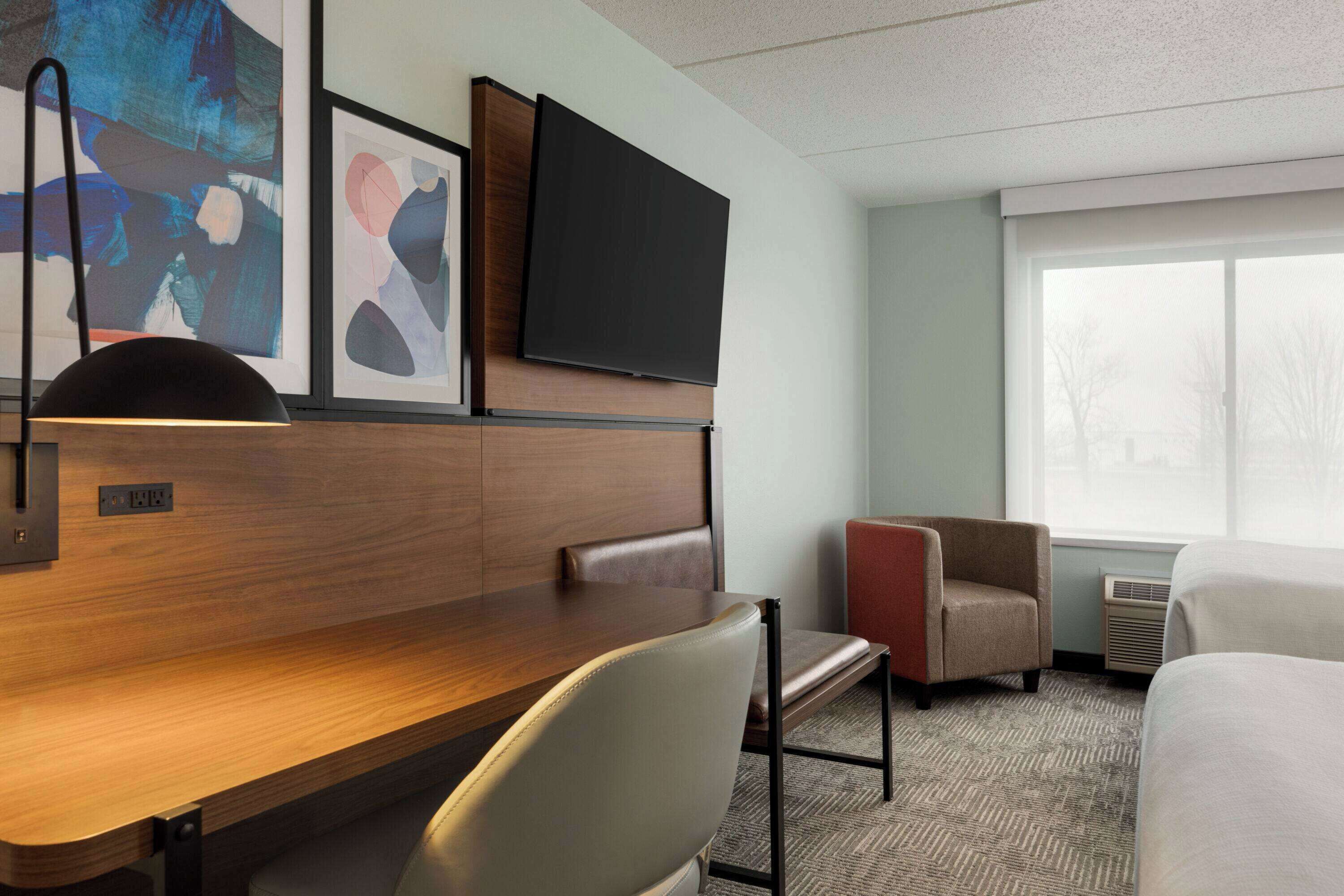 A hotel room with a wooden desk, a flat-screen TV mounted above it, a large window with a view, and two chairs.