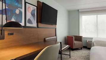 A hotel room with a wooden desk, a flat-screen TV mounted above it, a large window with a view, and two chairs.