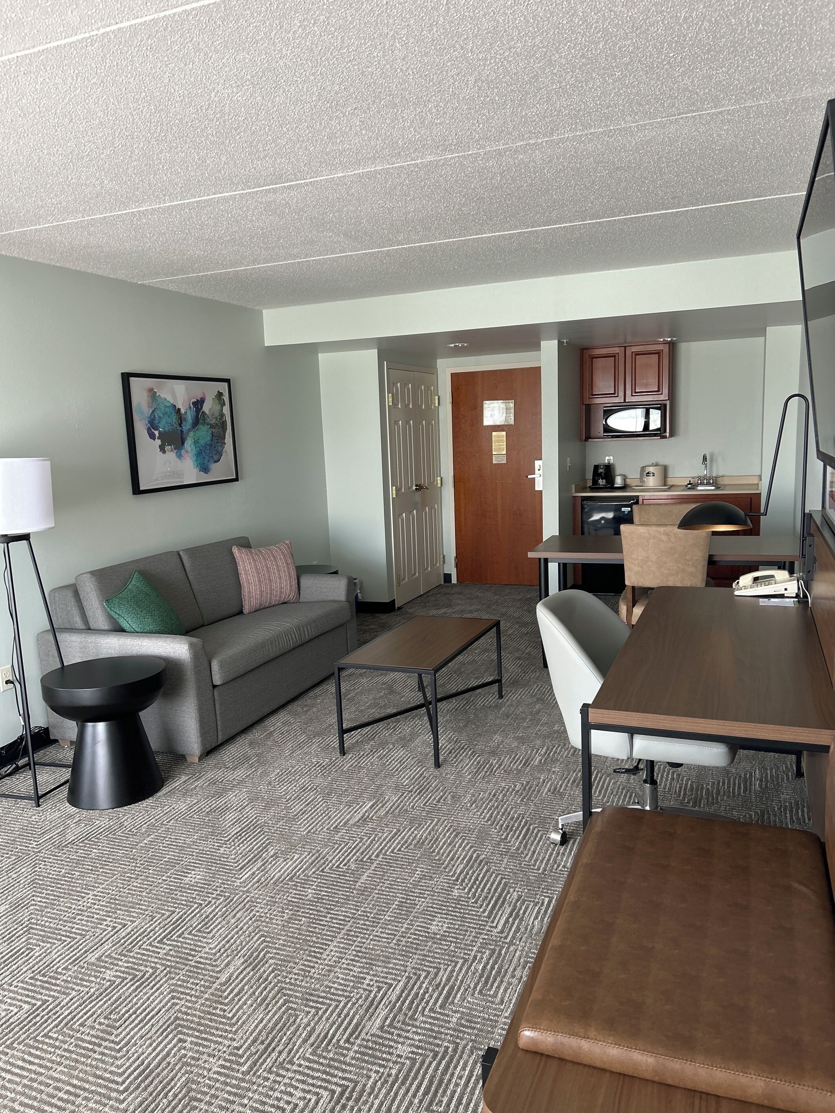 Ein modernes Hotelzimmer mit grauem Sofa, einem Couchtisch, einem kleinen Beistelltisch, einem Schreibtisch mit Stuhl und einer Küchenzeile.