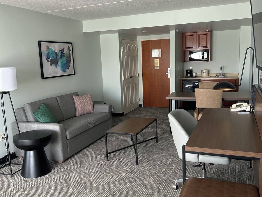 A modern hotel room with a grey sofa, a coffee table, a small side table, a desk with a chair, and a kitchenette area.