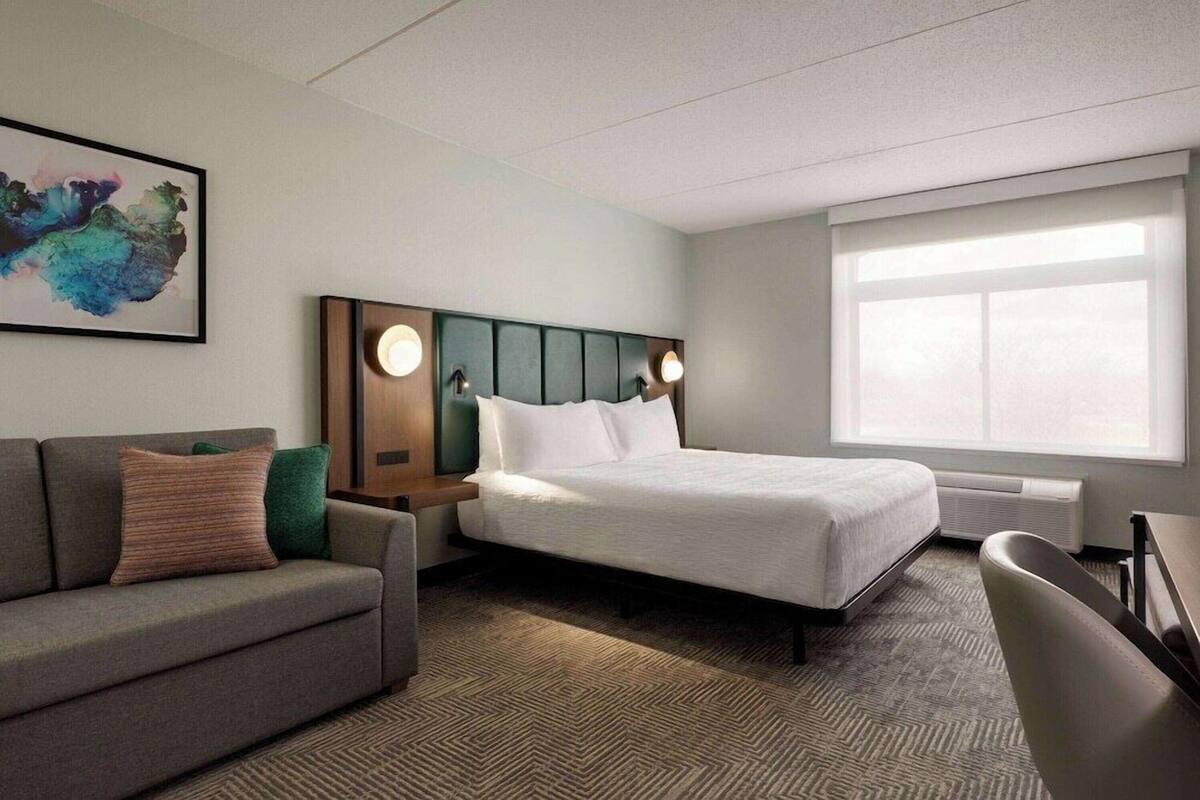 Habitación de hotel con cama, sofá, escritorio y un amplio ventanal.
