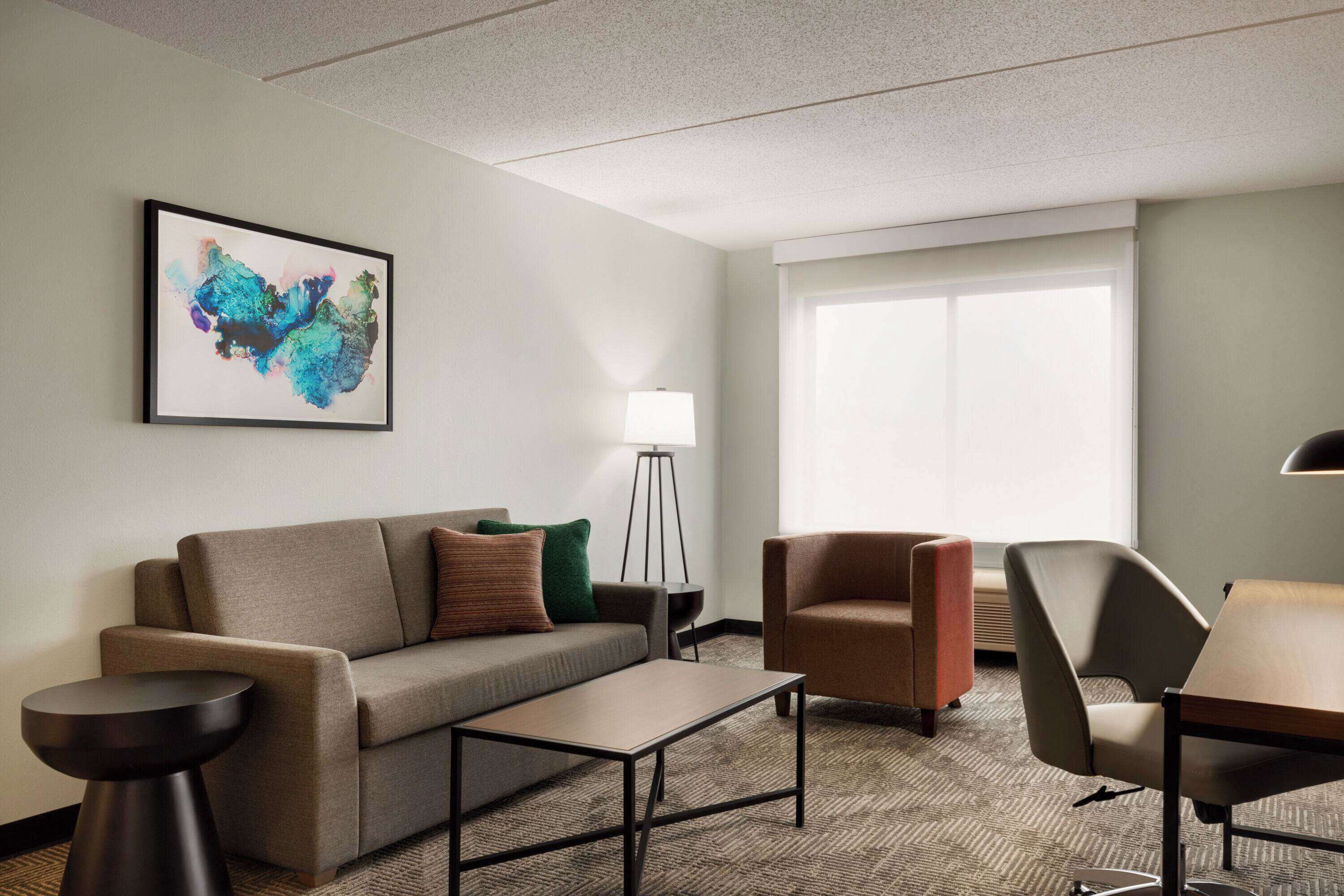 A modern hotel room with a sofa, armchairs, a small table, a floor lamp, and a large window.