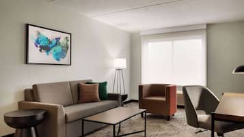 A modern hotel room with a sofa, armchairs, a small table, a floor lamp, and a large window.