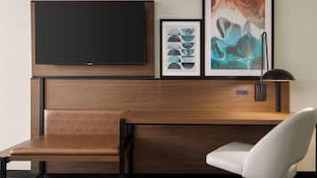 A modern hotel room with a wooden desk, a white chair, a flat-screen TV mounted above the desk, and two framed artworks on the wall.