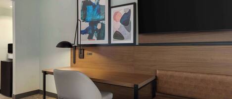 A hotel room with a wooden desk, a white chair, a sofa, and two framed abstract paintings on the wall.