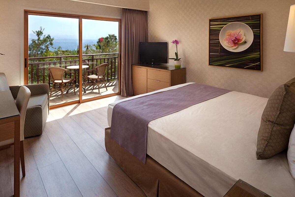 A hotel room with a bed, a desk, a chair, a television, and a balcony with a view of the ocean.