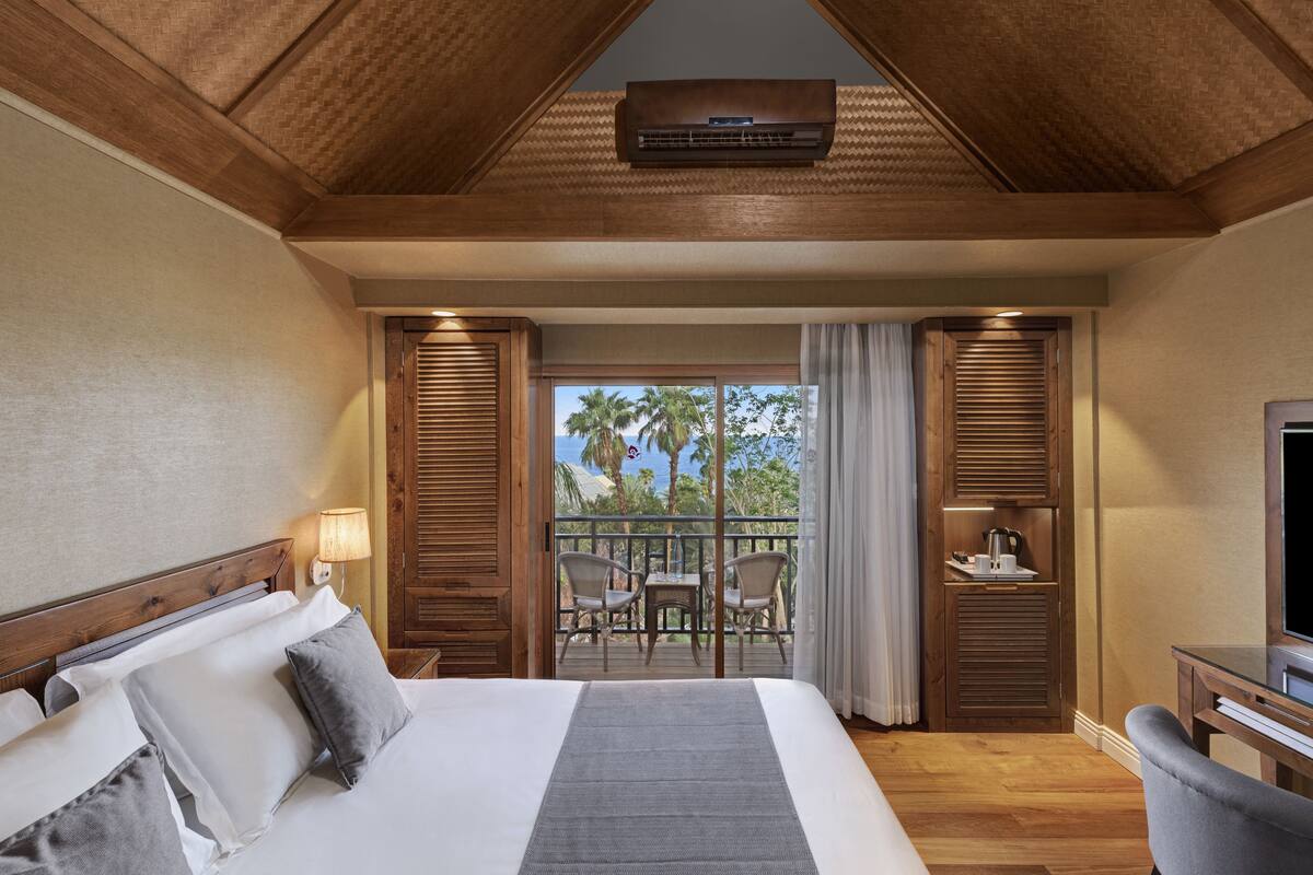 Habitación de hotel con una cama grande, televisión, balcón con vistas a palmeras y techo de madera.
