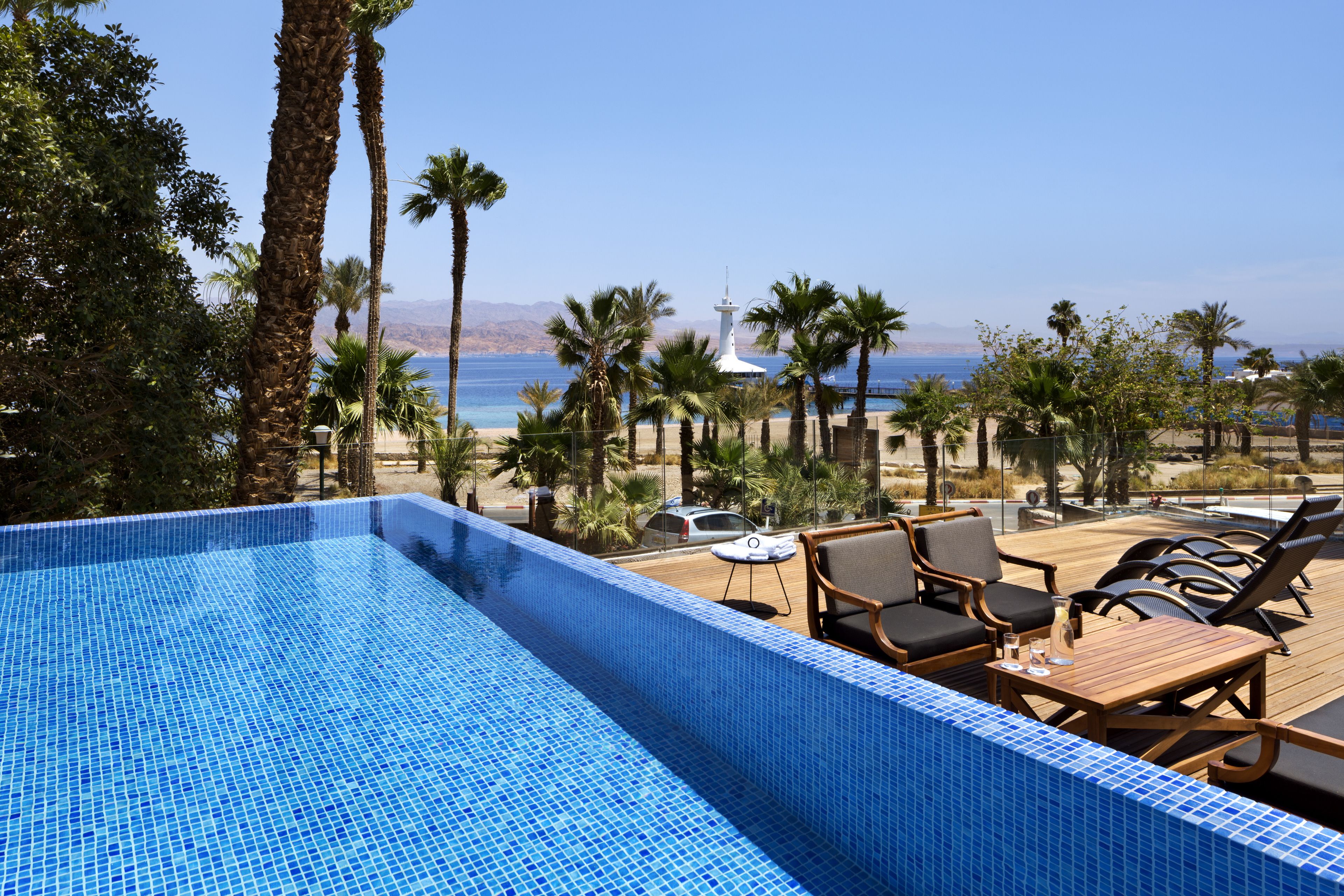 Zona de piscina con vista al mar, rodeada de palmeras y una terraza con sillones de descanso.
