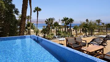Zona de piscina con vista al mar, rodeada de palmeras y una terraza con sillones de descanso.