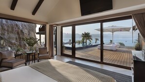 Ein geräumiges Schlafzimmer mit einem großen Bett, einer Sitzecke mit Stühlen, einem kleinen Tisch und Blick auf ein Schwimmbecken und Berge durch große Schiebetüren.