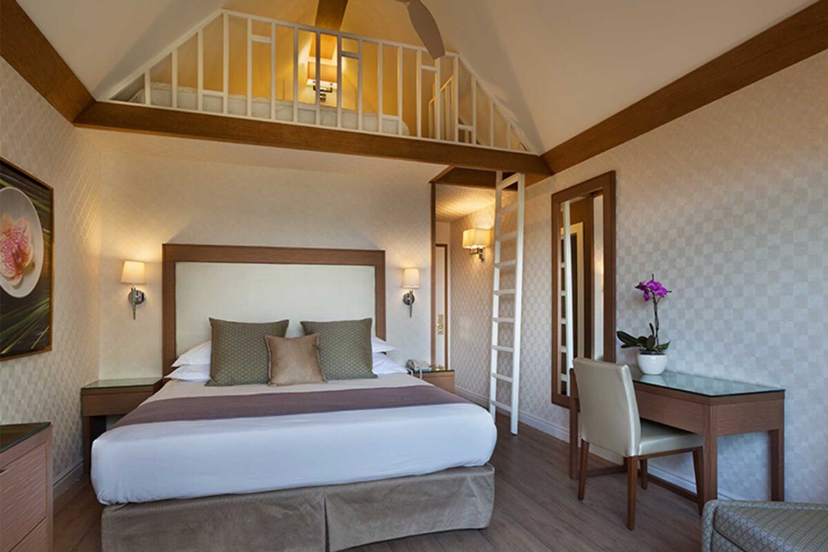A hotel room with a large bed, a desk, and a chair. There is a staircase leading to an upper level.