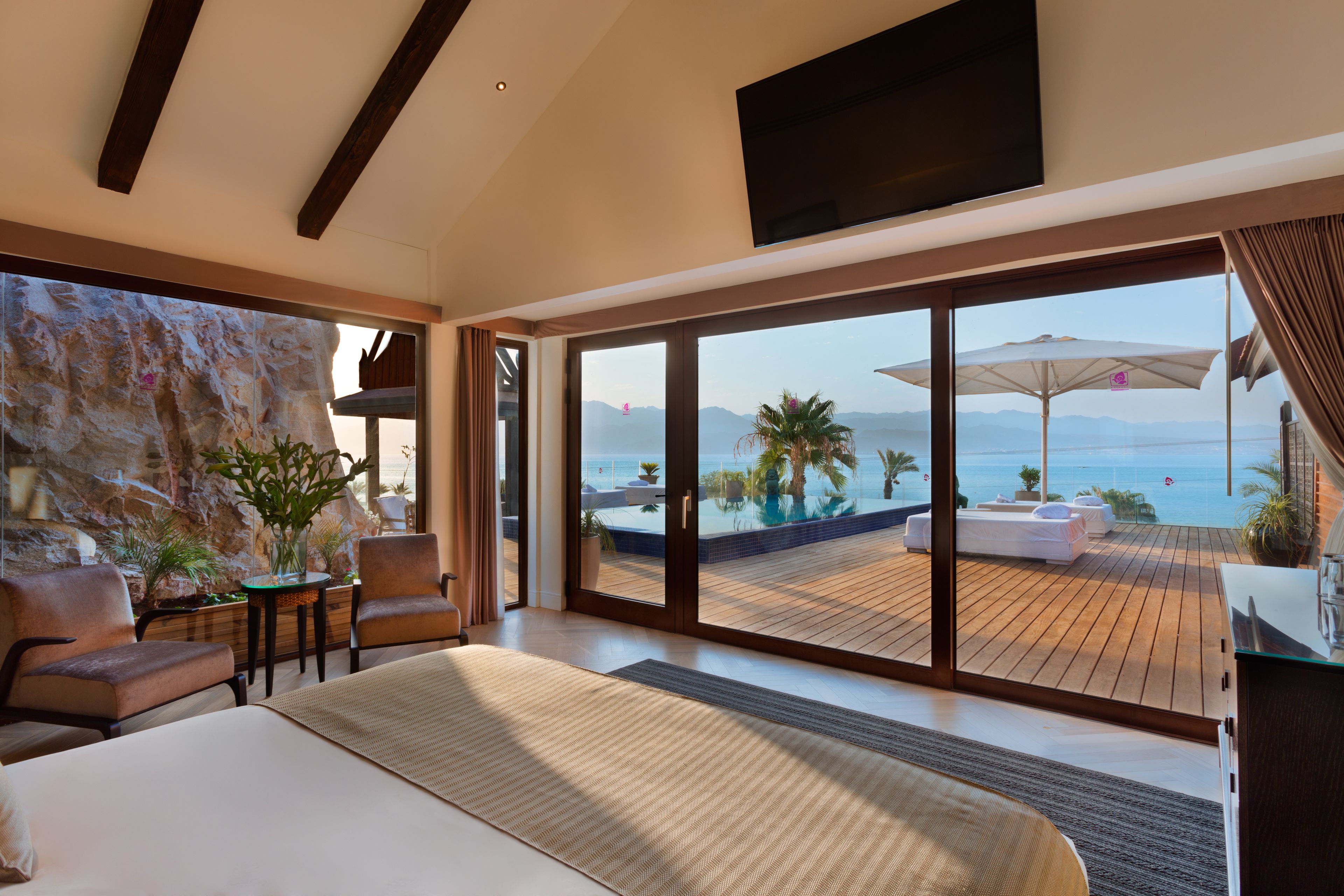 A modern bedroom with a large bed, a TV, and a view of a pool and mountains.