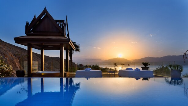 Ein ruhiger Infinity-Pool mit einer Holz-Laube und Liegestühlen, mit Blick auf einen Sonnenuntergang über Bergen.