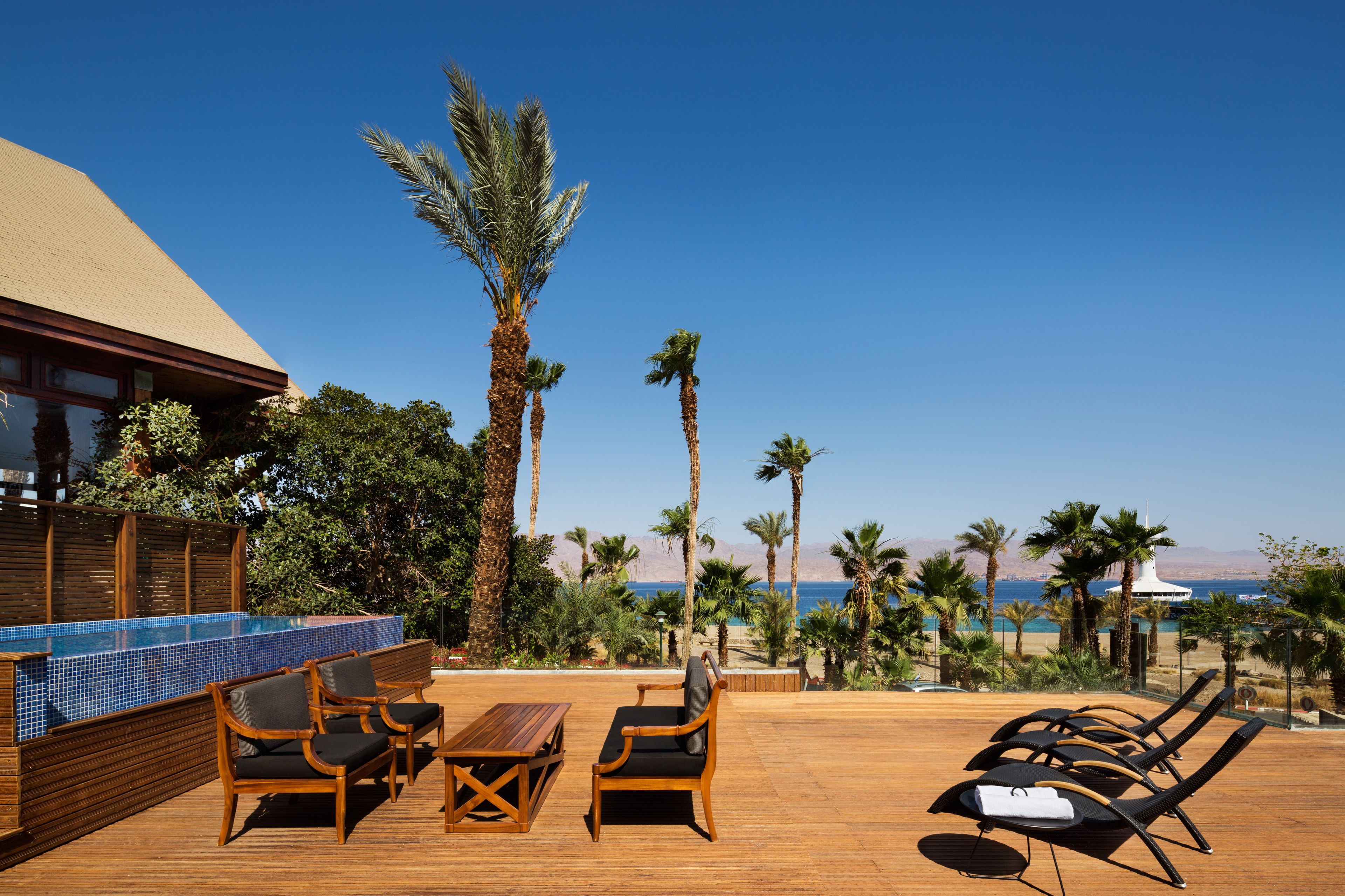 A wooden deck with lounge chairs and a pool area, overlooking a beach with palm trees and a clear sky.