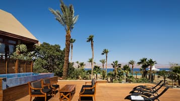 A wooden deck with lounge chairs and a pool area, overlooking a beach with palm trees and a clear sky.