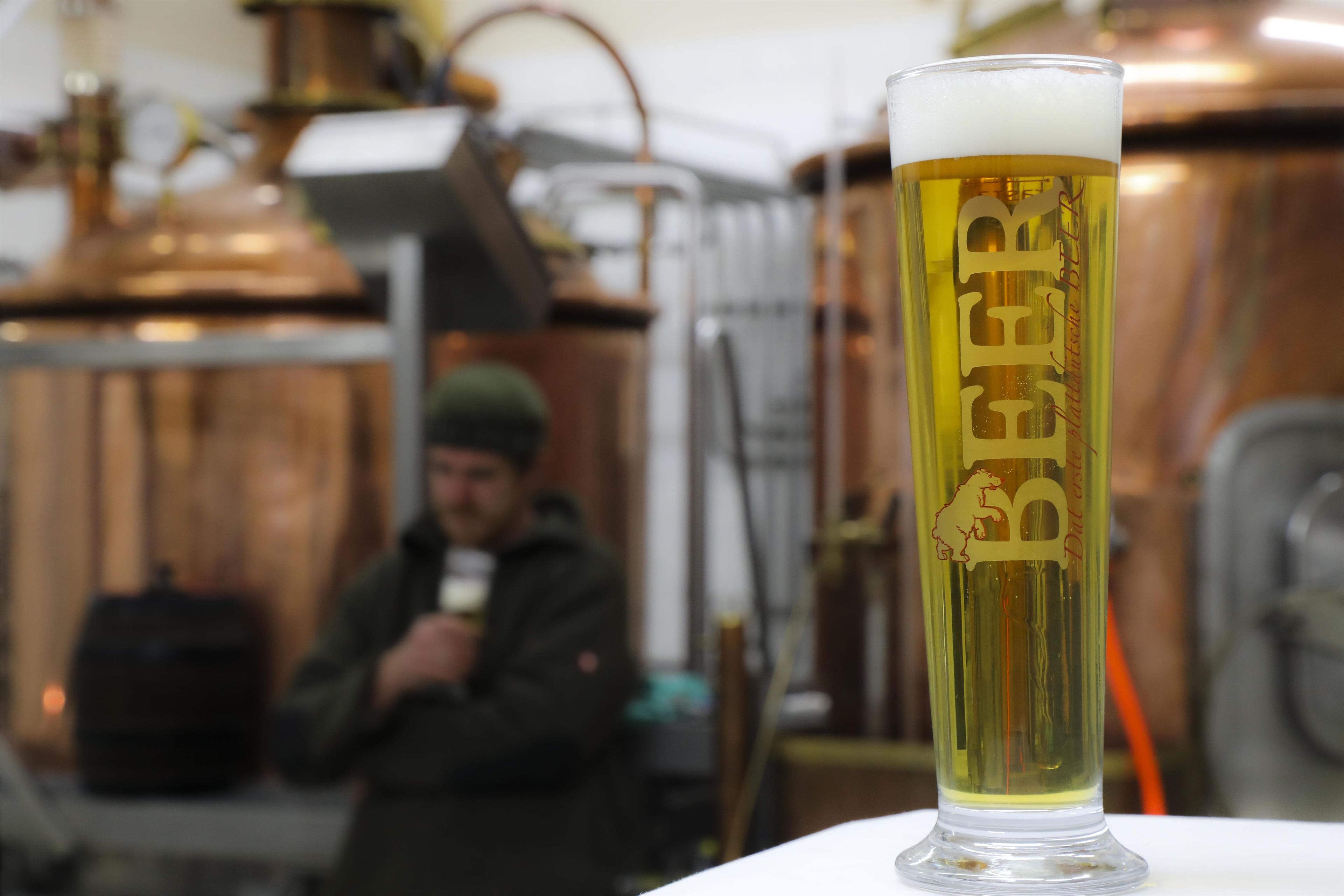 A glass of beer with a logo, placed in front of copper brewing equipment.