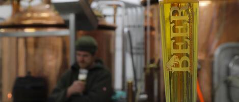 A glass of beer with a logo, placed in front of copper brewing equipment.