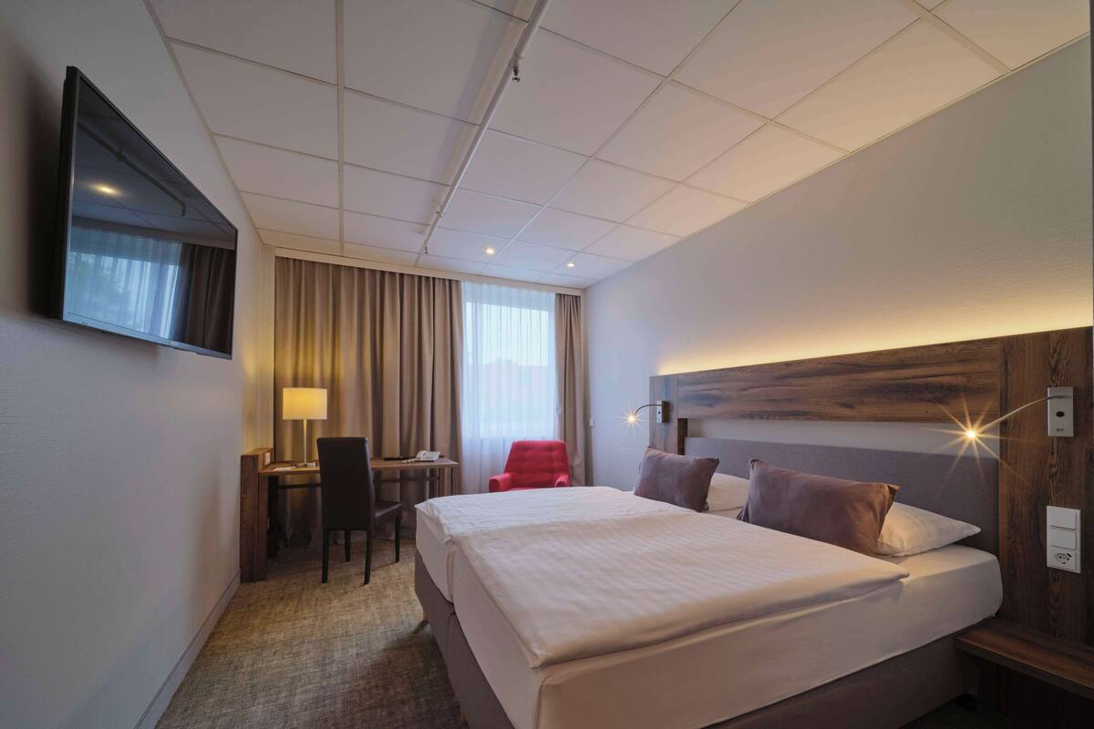 A hotel room with a large bed, a desk with a chair, a TV mounted on the wall, and a window with curtains.