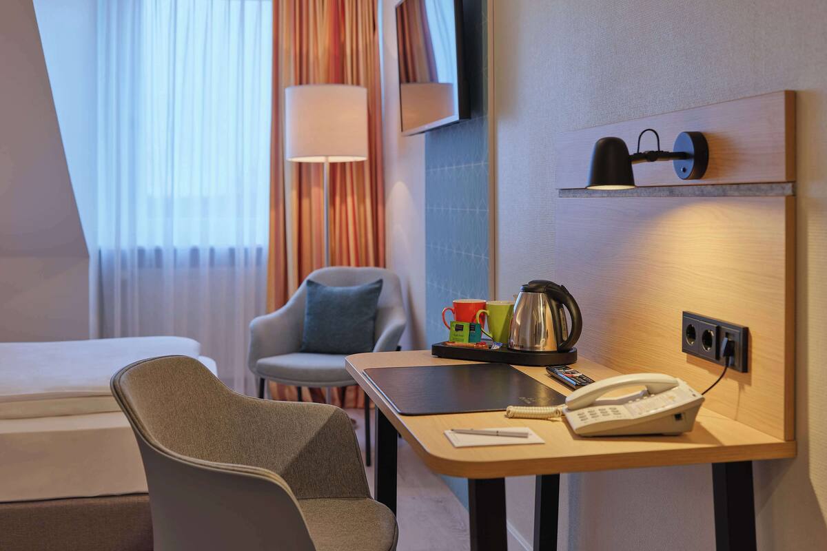 A hotel room with a bed, a desk with a telephone and a kettle, a chair, a TV mounted on the wall, and a window with curtains.