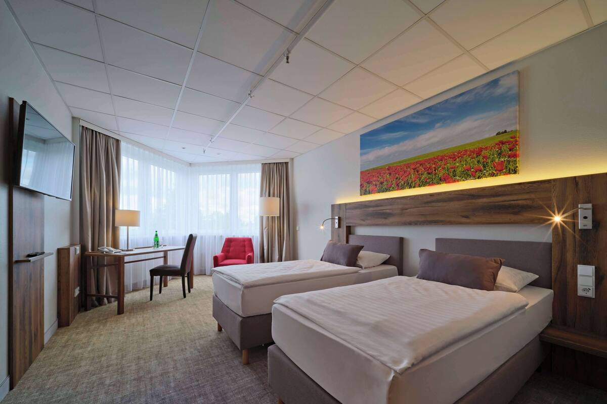 A hotel room with two beds, a desk with a chair, a TV, and a painting of a field.