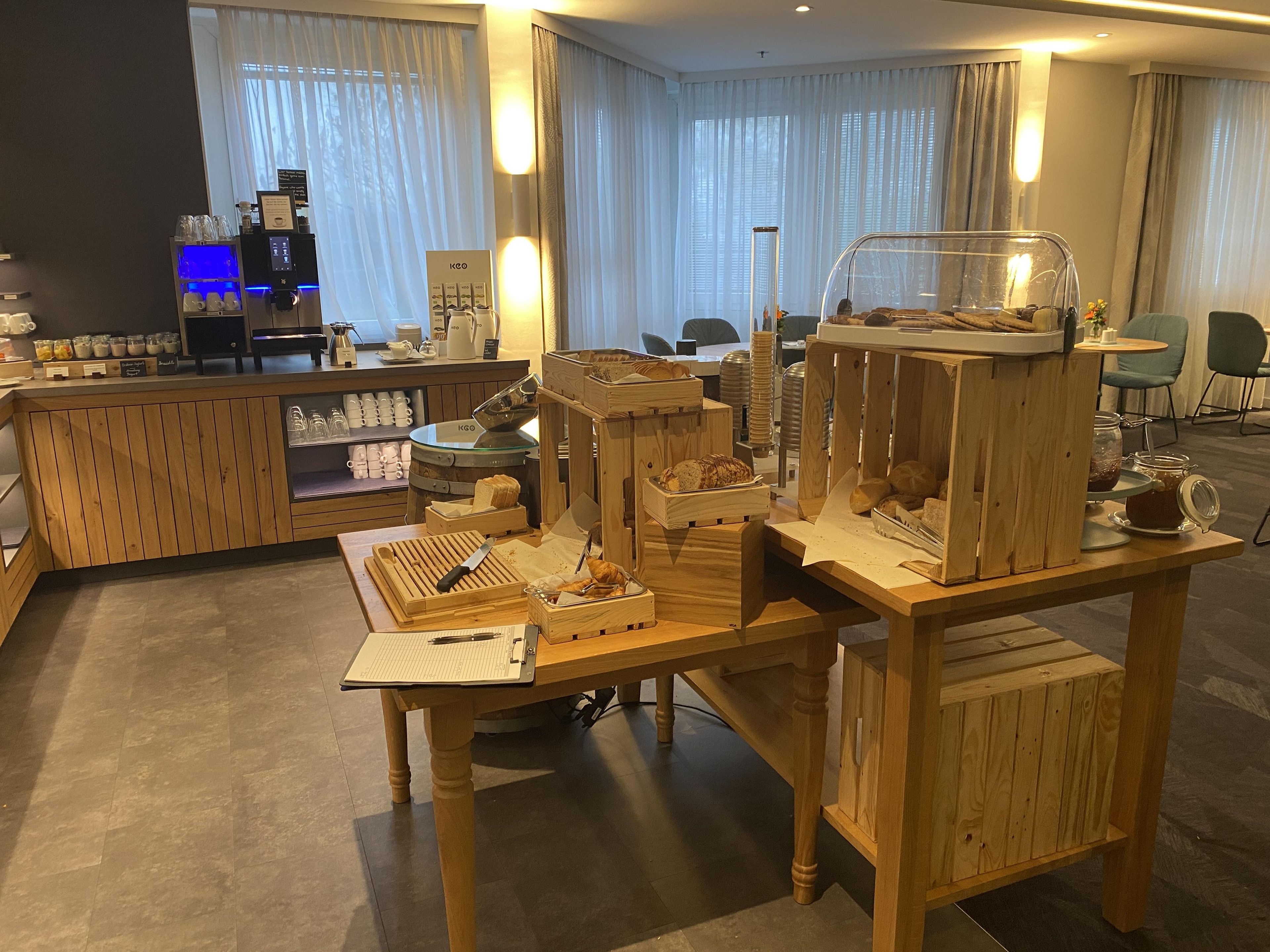 A breakfast buffet setup with wooden display cases, a coffee machine, and a seating area with chairs and a table.