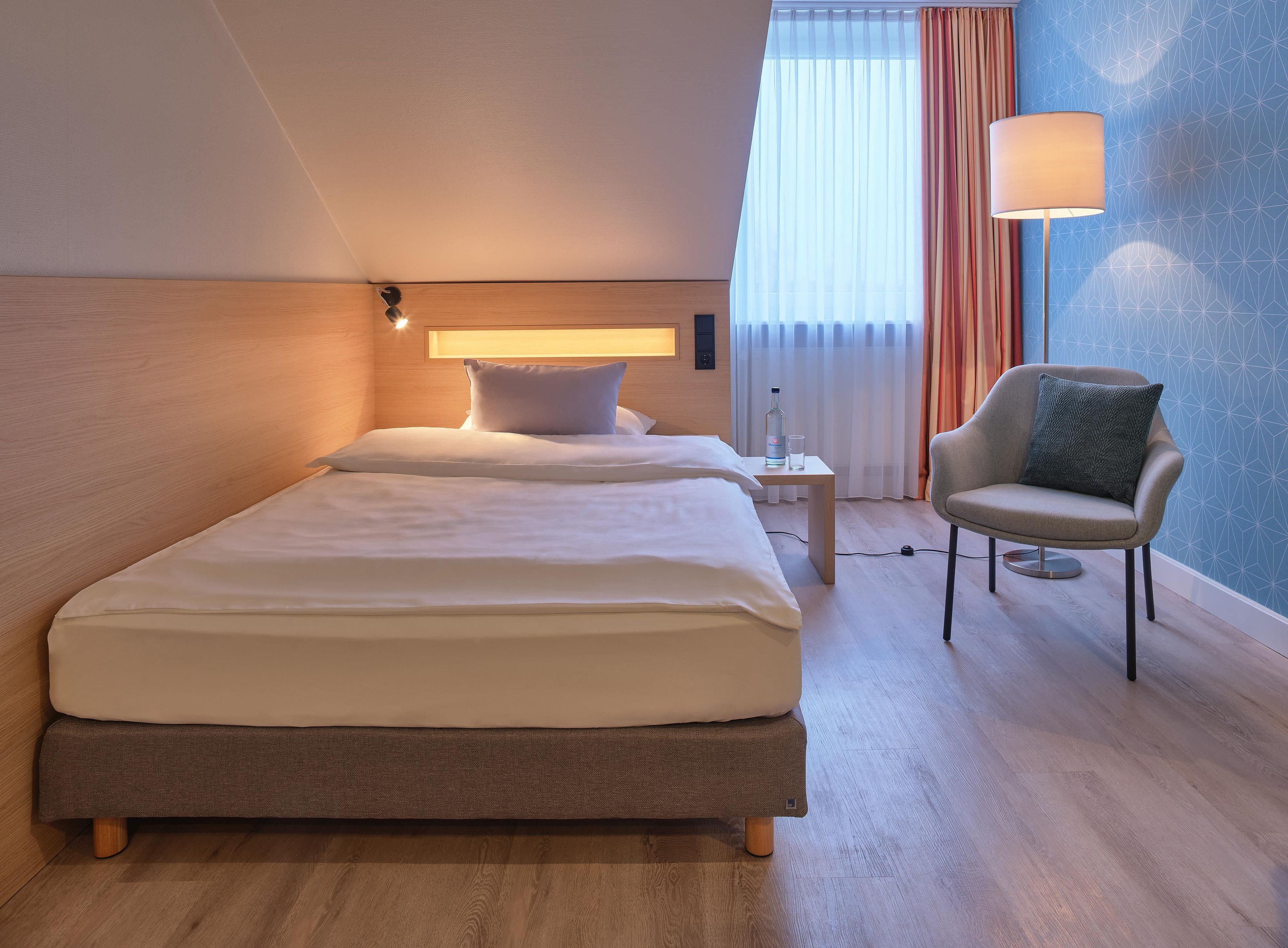 Una habitación de hotel con cama, mesita de noche, lámpara, silla y cortinas.